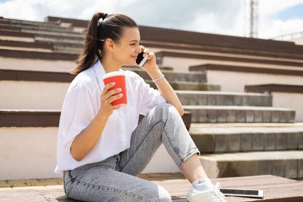 Happy teen girl talking on phone holding coffee paper cup enjoying sunny day sitting in amphitheater.