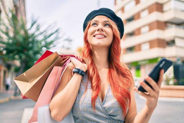 Young redhead girl holding shopping bags using smartphone at the city.