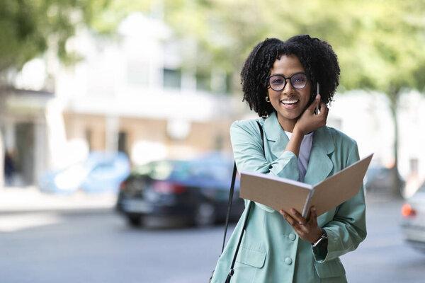 Portrait of cheerful pretty young black businesswoman with bushy hair wearing nice suit and eyeglasses walking by street and talking on phone, checking notes in her notepad, copy space