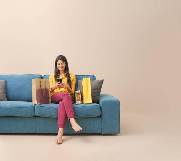 Happy young woman sitting on the couch with shopping bags and doing online shopping on her smartphone