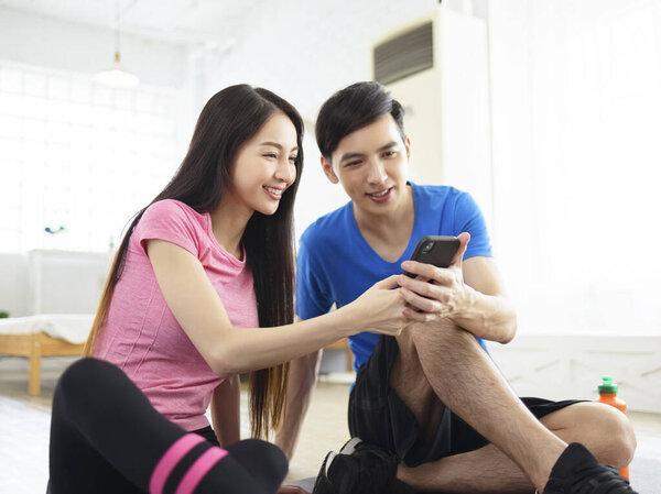 Athletic happy couple sitting on fitness mat, using smart phone.