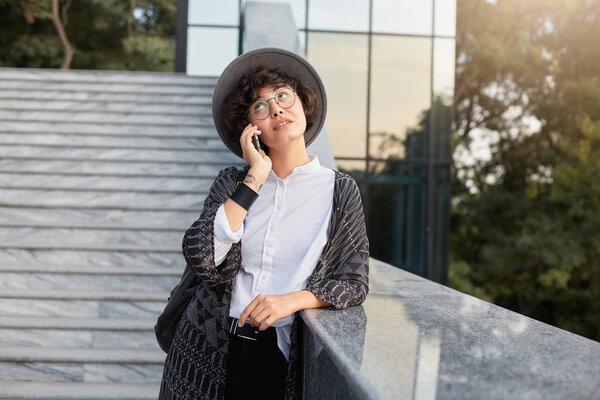 portrait of serious attractive young businesswoman talks on the phone and trying to correct her plans for a day, dressed casual, wears glasses and hat