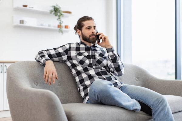 Joyful young man in casual clothes talking on smartphone while sitting on sofa in spacious dining room. Efficient Caucasian freelancer having business conversation with client of remote company.