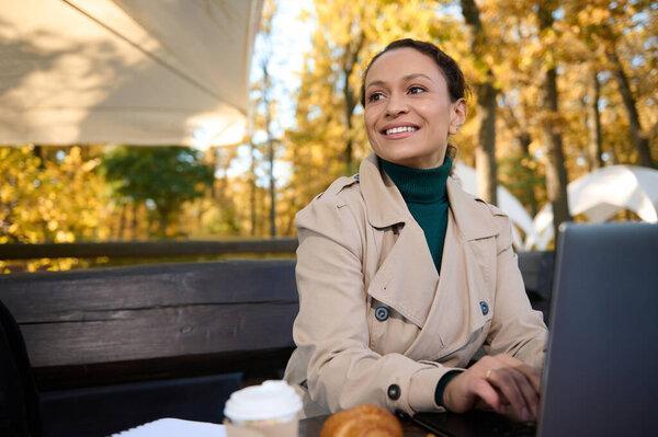 Charming business woman in trench coat sitting at the table of wooden cafeteria in the oak grove and looking the side, smiling with toothy smile while working on laptop on beautiful autumn background