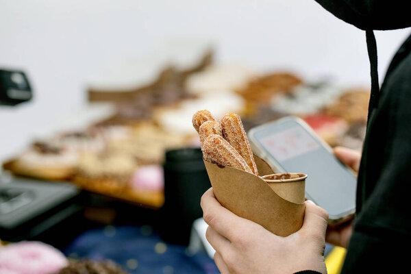 在食品市场上，人们用牛皮纸杯和智能手机拿着糖衣卷饼。夹杂着模糊烘焙柜台背景的甜甜的西班牙糕点.