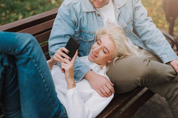 Top view of pretty blonde woman is using smartphone, having rest with her man on an alley bench. Romantic date