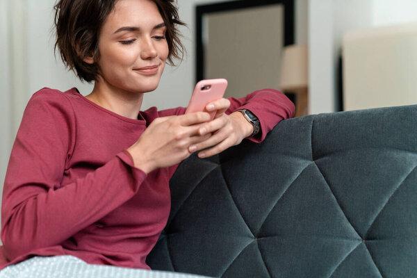 Photo of pleased young woman in leisure clothes using smartphone while sitting on sofa in living room