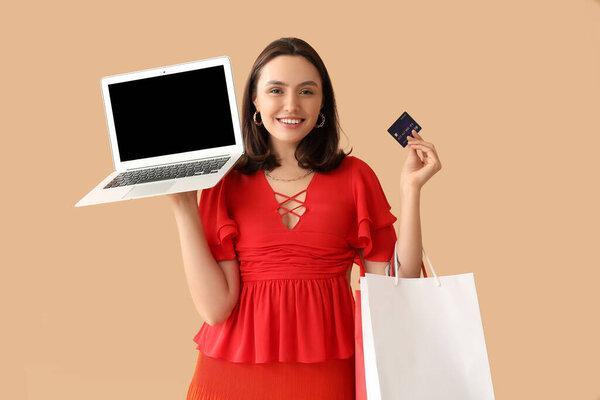 Young woman with credit card, laptop and shopping bags on beige background