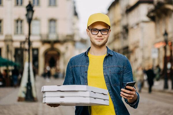 A young happy delivery man is making delivery of three pizzas. He is smiling. He is wearing a yellow cap and glasses. He is holding his phone and looking at the camera.