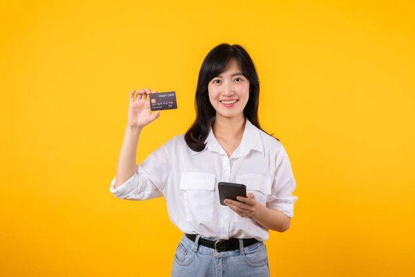形象年轻的亚洲女人穿着白衬衫，头戴牛仔，笑容满面，拿着信用卡，带着黄色背景的智能手机在网上购物。网上购物概念.