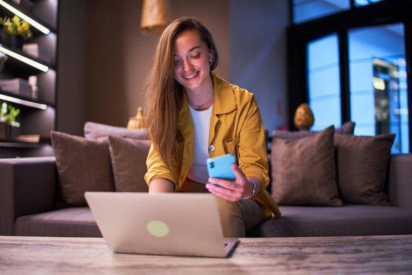 Happy cute joyful modern smiling satisfied smart young casual businesswoman wearing wireless headphones using laptop and smartphone for working online