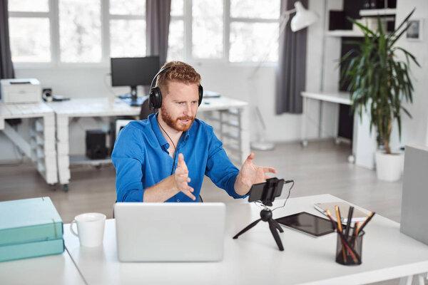 Young Caucasian employee having video call over smart phone while sitting in office. On head are headphones and on desk is laptop and coffee.