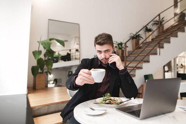 Bearded man in a suit sits in a cafe with a plate of salad and a laptop and offers a cup of coffee. Busy man in the cafe shows a cup of coffee in the camera.