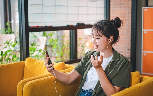 Smiling young asian woman using mobile phone checking social media,app playing game, shopping online, ordering delivery relax while sitting on a couch at cafe with laptop computer 