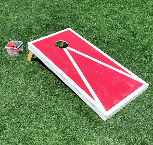 Red and white bean bag toss cornhole game on a green turf field.
