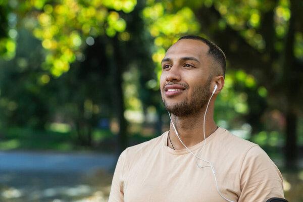 Close-up portrait of man in park, hispanic man wearing headphones listening to music and online audiobooks and podcasts, smiling and looking away, jogging and exercising outdoors.