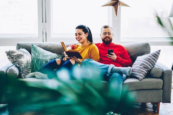 Cheerful hipster girl laughing on boyfriend's joke spending free time at cozy apartment together, positive woman reading book while her husband using smartphone for networking 