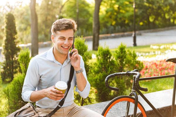 Happy young stylish man talking on mobile phone while drinking coffee outdoors with bicycle