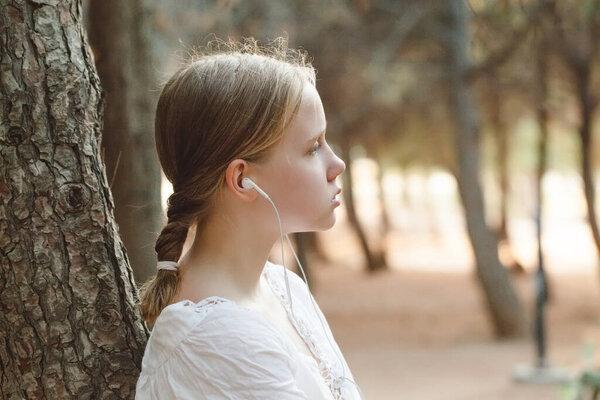 Porrait of pretty teenage girl in white shirt listen to music in headphones against forest background