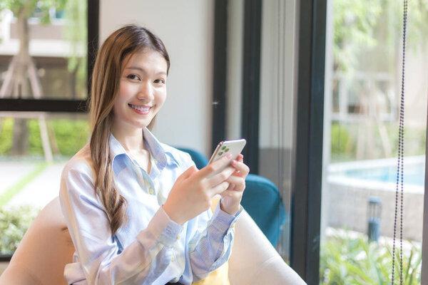 Asian beautiful woman with long hair who wear a long-sleeved shirt sits near the window while she uses smartphone to find information in working space.