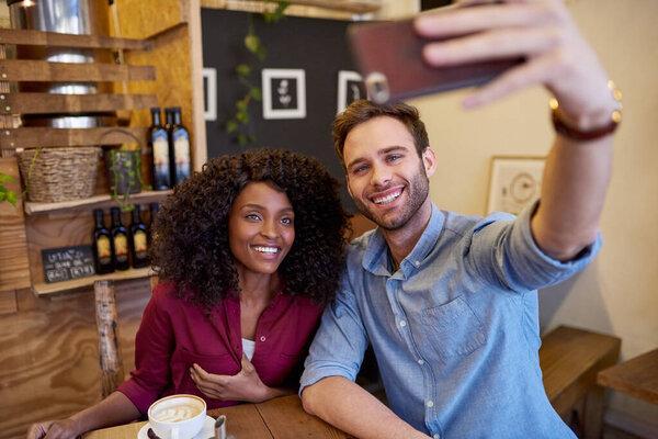 Diverse young couple smiling and taking selfie while sitting together at a table in a cafe