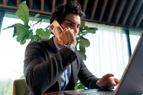 一位在办公室笔记本电脑上工作的商人用智能手机与同事交流的严肃男人的肖像