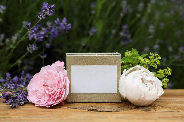 Paper package box. Blank label mockup for soap, facial cream. Rustic wooden table. Lavender herb, roses and alchemilla flowers. Blurred floral background. Skin product mockup scene, cosmetic product.