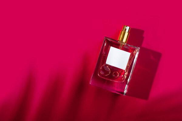 Transparent bottle of perfume with label on a red background. Fragrance presentation with daylight. Trending concept in natural materials palm leaves shadow. Womens and mens essence.
