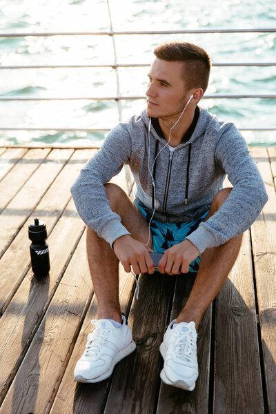 Concentrated sportsman in headphones, resting after workout, on pier.