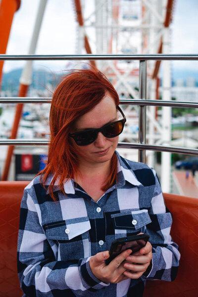 A woman is bored, she is not having fun on a stupid Ferris wheel attraction. Looking at smartphone. Entertainment and telecommunications concept. Vertical photo