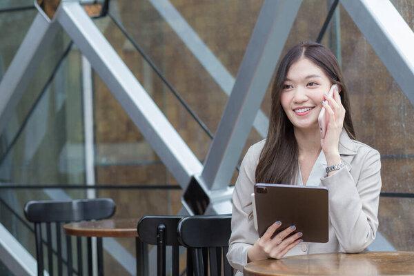 Beautiful young Asian professional business woman in a cream-colored suit holds a smartphone to talk, chat and record information happily on a tablet while in a coffee shop.
