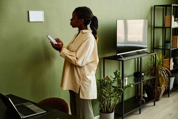 Portrait of young African American woman using smartphone while connecting to smart home system in modern interior